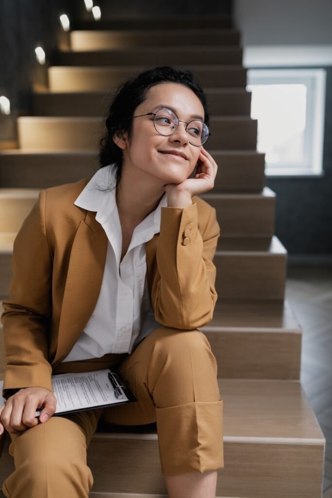 pexels-photo-7642125-7642125 Smiling businesswoman in glasses and corporate attire, sitting on staircase indoors with a clipboard.